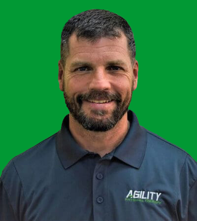 A man with a beard smiling in front of a green background, wearing a dark polo shirt with the logo "Alan Dalton Agility.