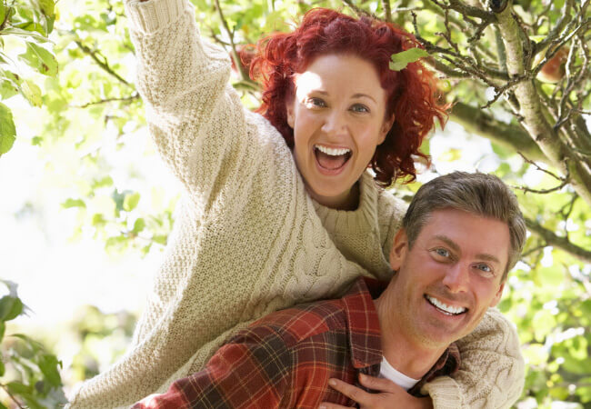 A joyful woman with red hair embracing a smiling man from the neck behind in an outdoor setting.