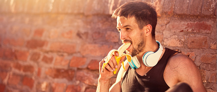 Man eating a banana while resting with headphones around his neck against a brick wall, taking a break from exercising to protect his joints.