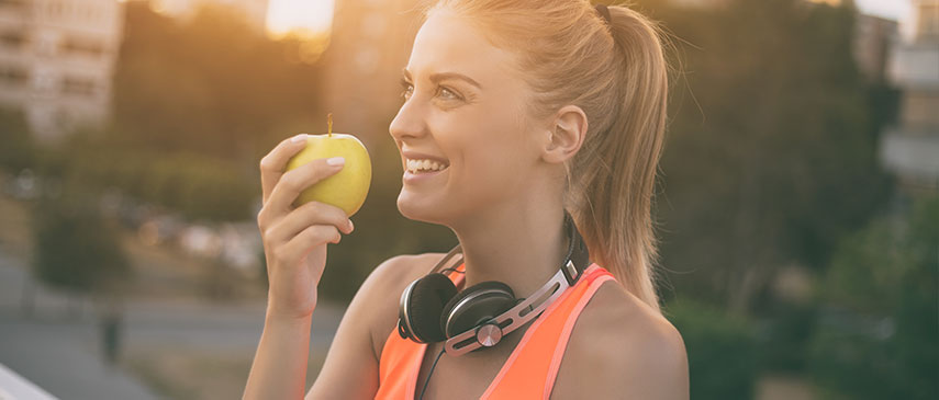 Woman smiling and holding an apple with headphones around her neck outdoors at sunset, her shoulder pain momentarily forgotten.