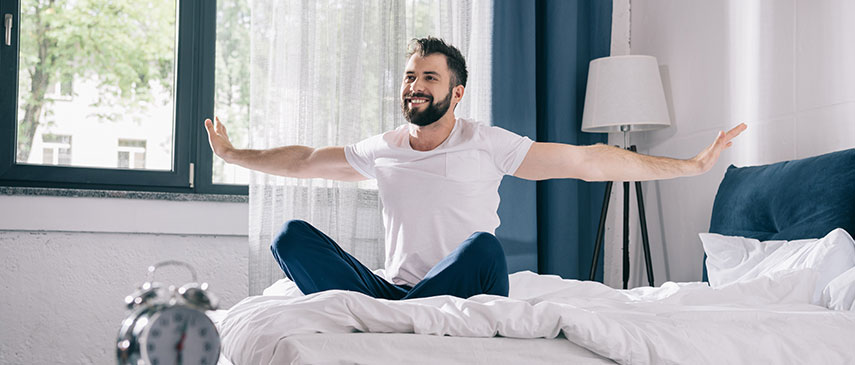 Man smiling and stretching his arms wide while sitting on a bed in a well-lit room, embracing his new morning routine that's set to change his life.