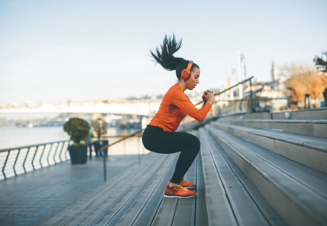 Woman experiencing knee pain while performing a squat exercise on steps outdoors.