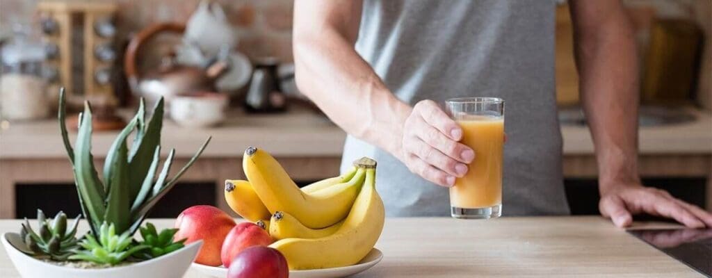 A person standing in a kitchen, holding a glass of juice as part of their nutritious diet, with fresh bananas and other fruits on the counter.