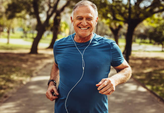 Senior man smiling and experiencing relief from aching necks while jogging with earphones in a park, enjoying better motion.