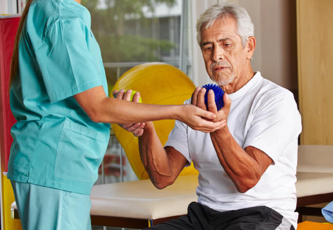 Senior man performing a rehabilitation exercise with a therapist using a stress ball to relieve arthritis pain naturally.