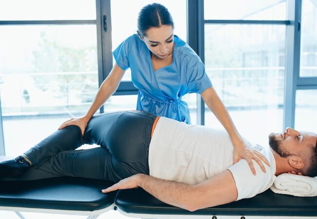 A physical therapist assisting a male patient with back pain by guiding him through a stretching exercise targeting his hips.