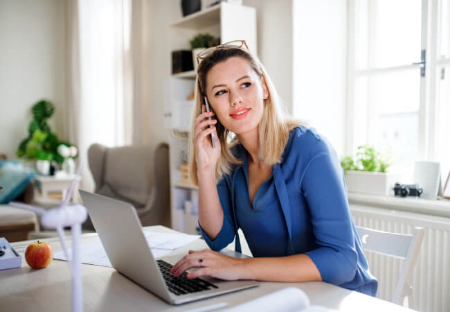 Woman in blue shirt talking on a telehealth appointment while working on a laptop in a bright office setting.