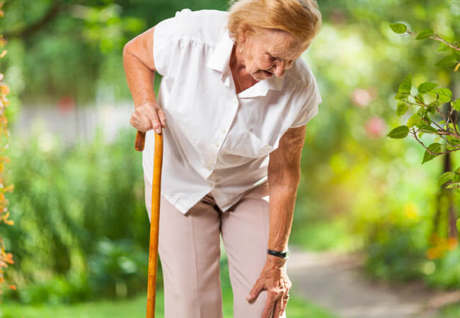 Elderly woman using a walking cane and experiencing knee pain outdoors seeks knee treatment.