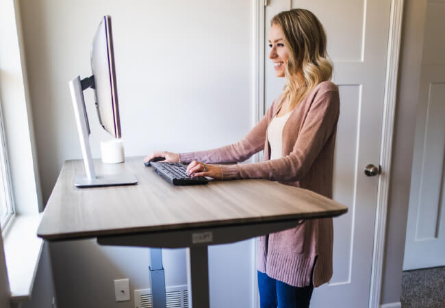 Woman standing at a standing desk working on a computer in a home office setting to avoid injuries.