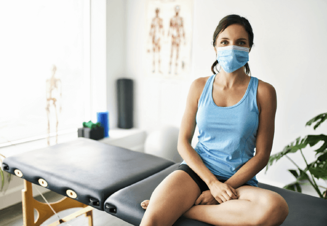 Woman in a mask sitting on an examination table in a medical office, seeking sciatica relief.