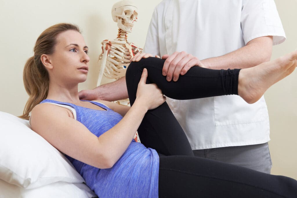 Physical therapist assisting a woman with knee pain through a leg exercise next to a skeletal model.