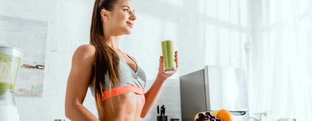 Woman in fitness attire holding a green smoothie in a physical therapy kitchen setting.