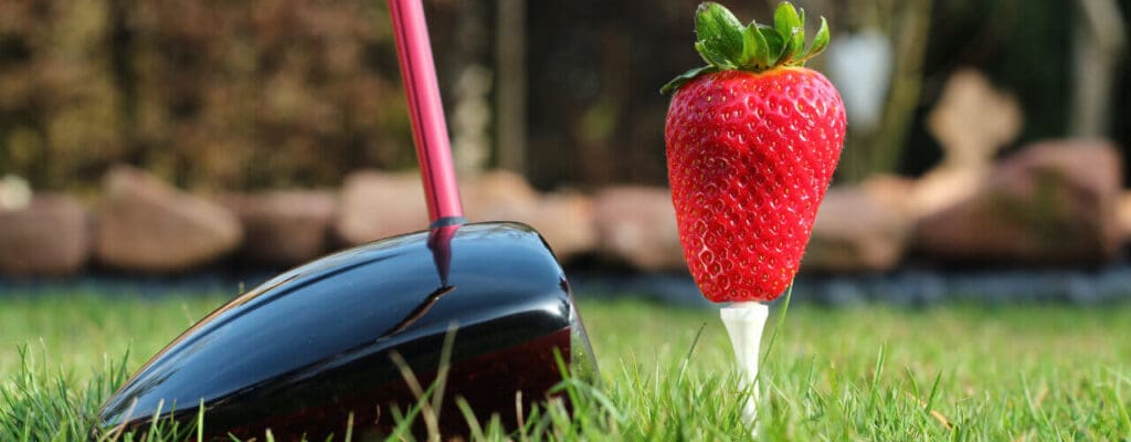 A giant strawberry placed on a golf tee at a golf course, with a golf club positioned behind it as if to take a swing.