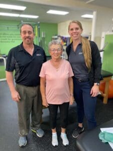 Three individuals in a physical therapy gym smiling at the camera: an elderly woman flanked by two professionals, possibly therapists, wearing company-branded attire. The individual on the right is Rebecca.