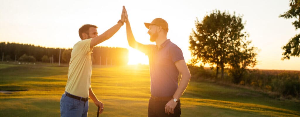 Two men engaging in a high-five outdoors during sunset, one visibly wincing from shoulder pain.