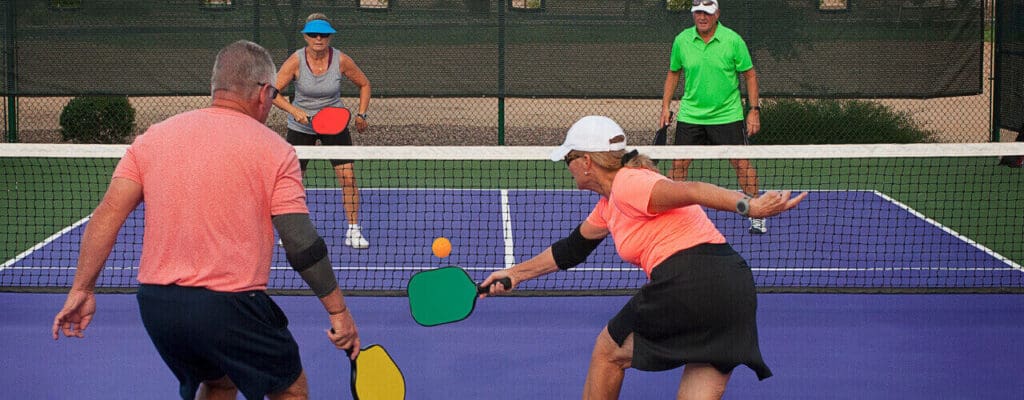 Four adults undergoing physical therapy playing a doubles match in pickleball, with one player reaching out to hit the yellow ball.