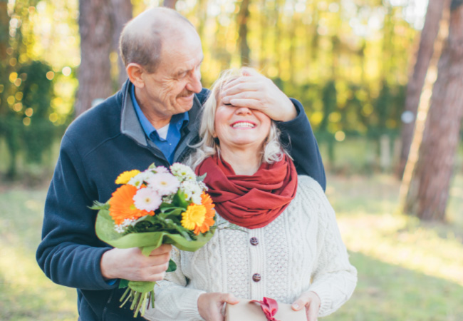 Senior man actively surprises woman with flowers, covering her eyes from behind in a park setting.