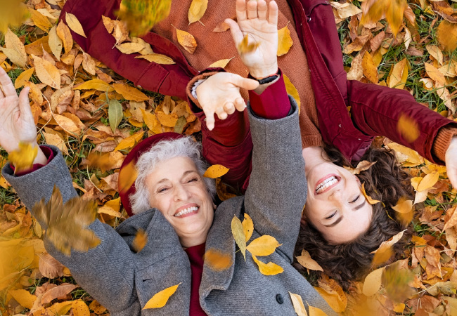 Two women lying on fall leaves, smiling and engaging in leaf prevention.