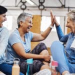 Three senior women sharing a joyful moment at a physical therapy session, with two of them high-fiving at a gym to help prevent injuries.