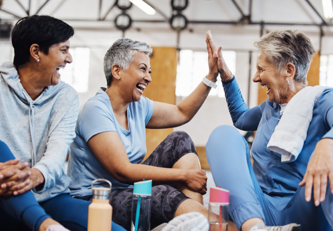 Three senior women sharing a joyful moment at a physical therapy session, with two of them high-fiving at a gym to help prevent injuries.
