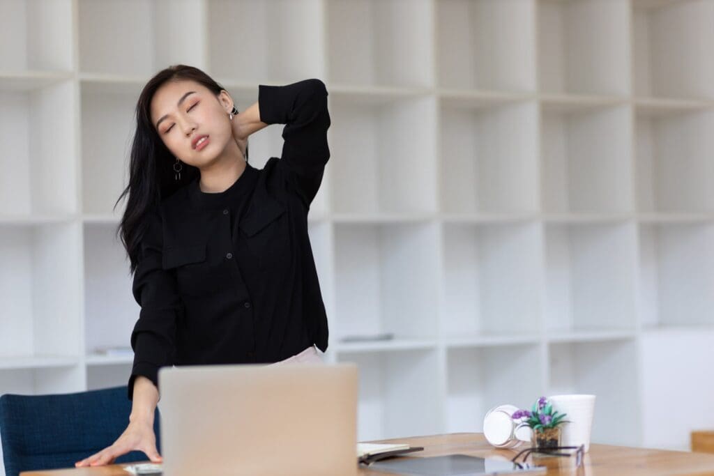 Woman stretching her neck while working at a laptop in an office setting to apply effective pain reduction methods for aches and pains.