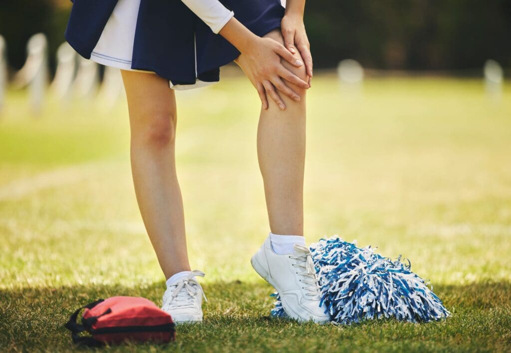Cheerleader holding knee in pain on the field with pom-poms and a bag of rehabilitation exercises on the ground.