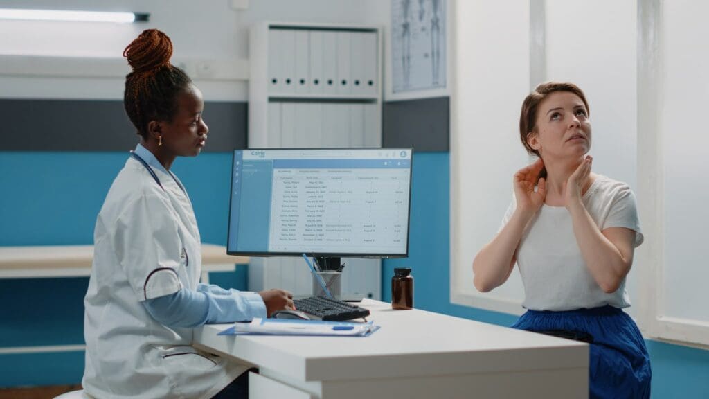 A healthcare professional is attending to a female patient who appears to be describing symptoms of neck pain and headaches while gesturing to her neck.