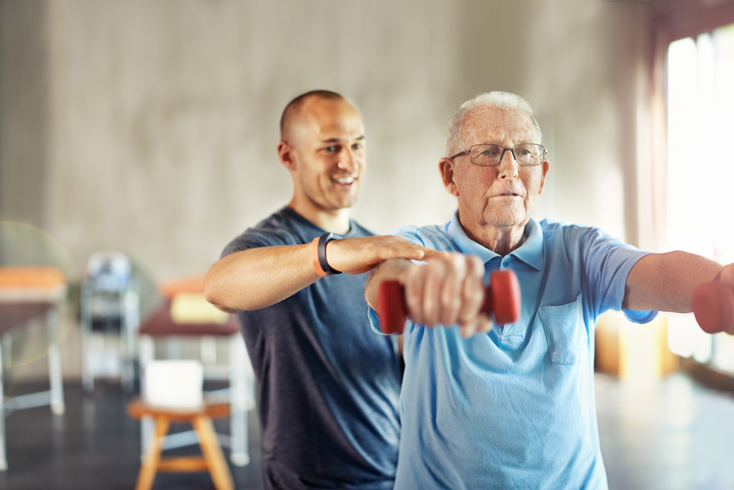 A senior man exercises with dumbbells, guided by a young male instructor in a fitness studio.