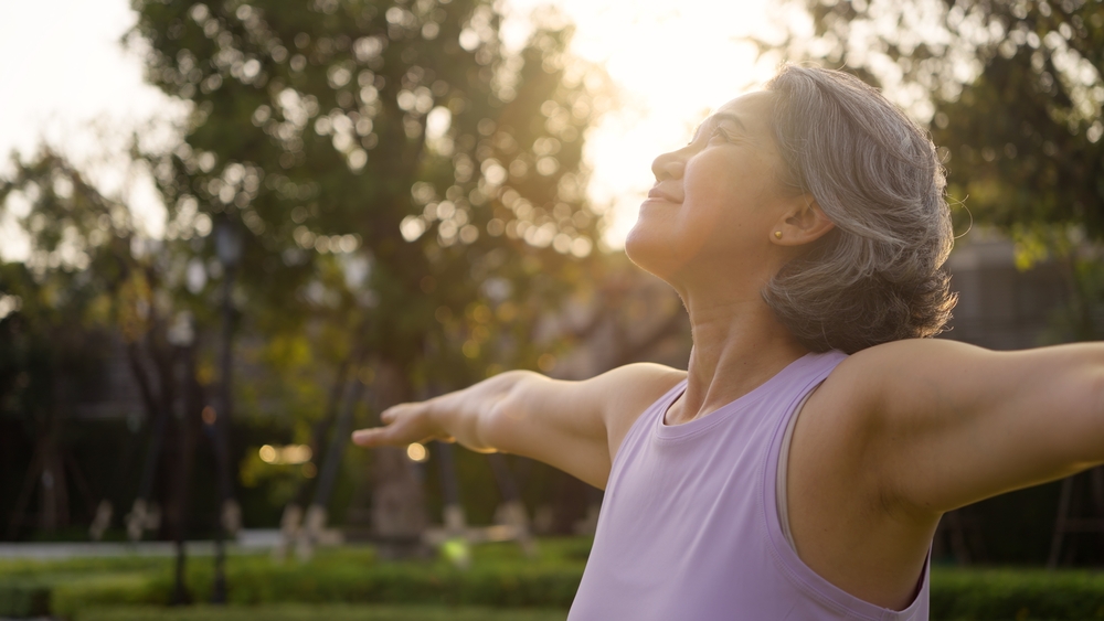 An older woman with gray hair stands outdoors with arms outstretched, smiling and facing the sunlight in a park setting, embodying the freedom and well-being often experienced after manual therapy.